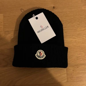 Svart Moncler mössa - Snygg svart ribbad mössa från Moncler med broderad logotyp framtill. Perfekt för att hålla värmen under kyliga dagar. Stilren design som passar till det mesta. Helt ny och skickas samma dag.