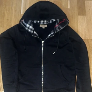 Svart huvtröja med dragkedja - Snygg svart huvtröja från Burberry med dragkedja och klassiskt rutigt mönster i luvan. Perfekt för en stilren och bekväm look. Tröjan har långa ärmar och praktiska fickor framtill.