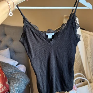 Svart ribbad linnetopp från Amisu - Säljer en svart ribbad linnetopp från Amisu med spetsdetaljer vid halsringningen. Toppen har smala axelband och en v-ringad design. Perfekt för lager-på-lager eller som den är.