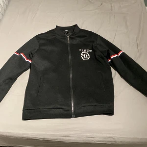 Svart jacka från Philipp Plein - Snygg svart jacka från Philipp Plein med dragkedja framtill. Jackan har vita och röda ränder på ärmarna och ett tryckt Philipp Plein märke på bröstet samt ryggen. Perfekt för en stilren look. Skriv för bättre bilder. Jackan har en liten spricka i armhålan men inget som syns eller märks.