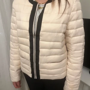 Beige pufferjacka med dragkedja - Säljer en snygg pufferjacka med svart dragkedja och detaljer. Jackan är varm beige och inte kommer till användning lägre. 
