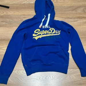 Blå hoodie från Superdry - Säljer en snygg blå hoodie från Superdry med gul text på framsidan. Den har en klassisk känguruficka och justerbar huva med vita snören. Perfekt för en avslappnad stil.