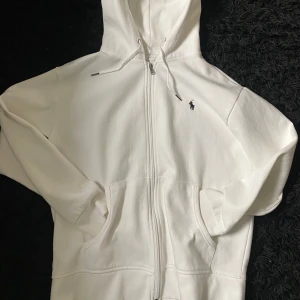Vit hoodie med dragkedja från Ralph Lauren - Säljer en stilren vit hoodie från Ralph Lauren med dragkedja och klassisk logga på bröstet. Den har en bekväm passform och är perfekt för en avslappnad stil. Huvan har snörning och det finns praktiska fickor framtill.
