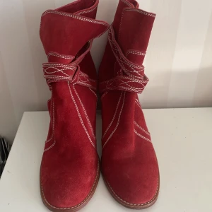 Whyred Röda mocka boots med klack - Snygga röda boots från Whyred  i mocka med dekorativa vita sömmar och en stabil klack (7 cm) . Skorna har en unik design med remmar som går runt ankeln och en rundad tå. Perfekta för att ge en färgklick till din outfit. Provade och ej använda, så i nyskick!
