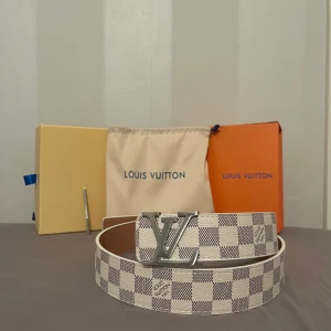Rutig Louis Vuitton bälte - Snyggt Louis Vuitton bälte med klassiskt rutigt mönster i beige och vit. Bältet har ett stort LV-spänne i silverfärg som ger en lyxig touch. Perfekt för att lyfta vilken outfit som helst.