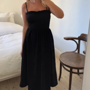 Svart axelbandslös klänning - Säljer en stilren svart axelbandslös klänning med smala axelband. Klänningen har en smockad överdel som ger en bekväm passform och en luftig kjol som faller fint. Perfekt för varma dagar eller kvällar. Jättesnygg 
