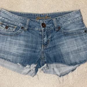 Blå jeansshorts från Guess - Snygga blå jeansshorts från Guess med fransiga kanter och klassisk femficksdesign. Perfekta för en avslappnad sommarlook. Shortsen har en knapp och dragkedja framtill samt bälteshällor.