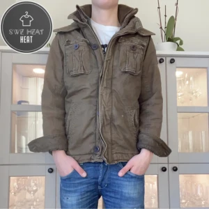 Olivgrön jacka från Abercrombie & Fitch - Säljer en olivgrön jacka från Abercrombie & Fitch med huva och flera fickor. Jackan har knappar och dragkedja framtill samt ribbade detaljer vid kragen. Perfekt för kyligare dagar.