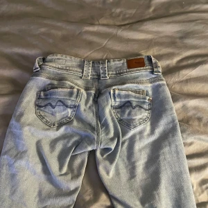 Ljusblå jeans med låg midja - Säljer ett par snygga ljusblå jeans med låg midja och straight fit. De har en sliten look med små revor och två knappar i midjan. Bakfickorna har ett enkelt mönster. Perfekta för en avslappnad stil. Stl 25,30