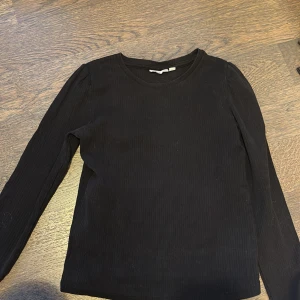 Svart ribbad tröja från KappAhl - Säljer en stilren svart ribbad tröja från KappAhl. Tröjan har långa ärmar och en rund halsringning, perfekt för en enkel och snygg look. Passar bra till både jeans och kjol.