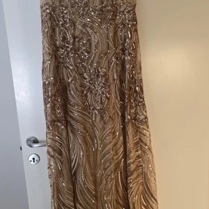 Champagnefärgad paljettklänning - Elegant champagnefärgad långklänning med glittrande paljetter och intrikata mönster. Klänningen har en snörning i ryggen och är axelbandslös, vilket ger en sofistikerad look. Perfekt för speciella tillfällen.