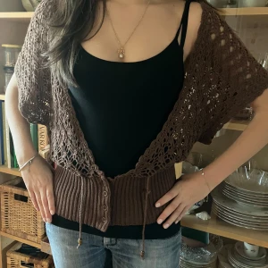 Brun stickad bolerotopp - Säljer en brun stickad bolerotopp med ett vackert hålmönster. Toppen har korta ärmar och knappar framtill. Perfekt för att lägga till en bohemisk touch till din outfit. Passar bra över en enkel topp eller klänning.