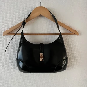 Svart axelväska i glansigt skinn - Snygg svart axelväska i skinn med stilren design. Väskan har en justerbar axelrem och ett elegant spänne framtill. Perfekt för att ge en klassisk touch till din outfit.