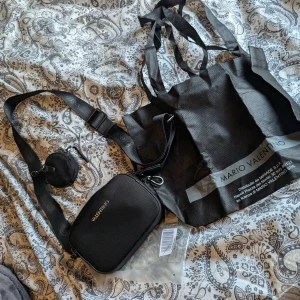 Svart axelväska från Mario Valentino - Snygg svart axelväska från Mario Valentino med justerbar axelrem och dragkedja. Väskan har en minimalistisk design och inkluderar en rund liten väska som kan fästas på remmen. Perfekt för att bära dina nödvändigheter med stil. Enbart används en gång. 