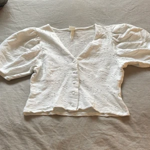 Vit blus från H&M - Säljer en söt vit blus från H&M med puffiga kortärmar och knappar framtill. Blusen har en v-ringad design och ett diskret mönster i tyget. Perfekt för en stilren look! 💗
