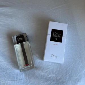 Dior Homme Sport Parfym - Säljer en elegant Dior Homme Sport parfym. Flaskan är stilren och rektangulär med en svart detalj i mitten. Förpackningen är vit med svart och röd text. Perfekt för den som söker en fräsch och sportig doft.