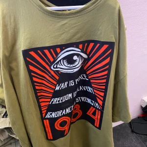 Olivgrön t-shirt med 1984 tryck - Säljer en olivgrön t-shirt med ett stort, iögonfallande tryck i svart, rött och vitt. Trycket har ett öga och texten 'War is Peace, Freedom is Slavery, Ignorance is Strength, 1984'. Perfekt för den som gillar unika och uttrycksfulla plagg.