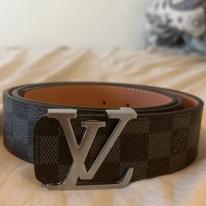 Rutigt bälte från Luviton - Snyggt bälte från Luviton med ett  rutigt mönster i svart och grått. Bältet har en stor Luviton-logotyp i silver som spänne. Perfekt för att göra dig se stilig och lyxig ut. Otroligt snygga till gråa jeans men funkar på alla såklart. Känns bra och är bekväm att ha på sig. Serveras med en 4cm metallpinne. Nästan helt oanvänd och jätte fräsch. PRISET KAN DISKUTERAS