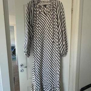 Randig långklänning med knytband - Säljer en stilren långklänning med svartvita ränder och knytband vid halsen. Klänningen har långa ärmar och en luftig passform, perfekt för en avslappnad look. Den är gjord i ett lätt och bekvämt material.