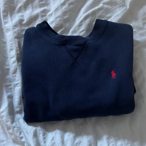 Mörkblå tröja från Ralph Lauren - Säljer en stilren mörkblå tröja från Ralph Lauren med det klassiska röda logotypen på bröstet. Tröjan har en rund halsringning och är perfekt för en avslappnad stil. Passar bra till både jeans och chinos.