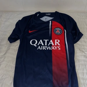 Blå fotbollströja från Nike - Säljer en blå fotbollströja från Nike med röd och vit detalj. Tröjan har korta ärmar och klubbmärke på bröstet. Perfekt för fotbollsfans!