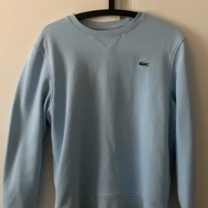 Ljusblå sweatshirt från Lacoste - Säljer en stilren ljusblå sweatshirt från Lacoste med det klassiska krokodilmärket på bröstet. Tröjan har långa ärmar och en rund halsringning. Perfekt för en avslappnad look. 