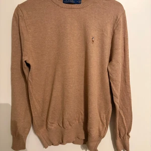 Beige tröja från Polo Ralph Lauren - Säljer en stilren beige tröja från Polo Ralph Lauren i mjuk pima bomull. Tröjan har en broderad logotyp på bröstet och ribbade muddar vid ärmslut och nederkant. Perfekt för en avslappnad och elegant look.
