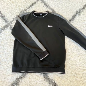 Sweatshirt från Boss - Snygg svart Sweatshirt från Boss med grå vita detaljer längs ärmarna och vit text på bröstet. Perfekt för en stilren look. Tröjan har långa ärmar och rund halsringning.                                                            OBS…  Storleken är ”L” men passar ”M”.