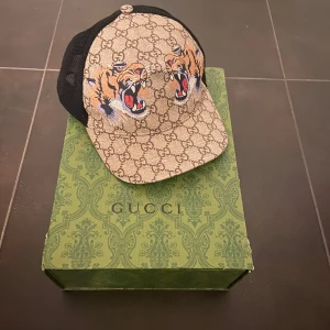Gucci keps med tigerbroderi - Snygg keps från Gucci med ikoniskt GG-mönster och broderade tigrar framtill. Kepsen har ett justerbart band baktill i grönt och rött. Perfekt för att ge din outfit en lyxig touch.