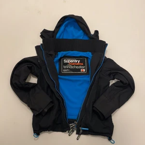 Svart vindjacka från Superdry - Säljer en svart vindjacka från Superdry med blå detaljer och broderad logga. Jackan har en huva och dragkedja framtill. Insidan är fodrad med blått tyg för extra komfort. Perfekt för blåsiga dagar! 