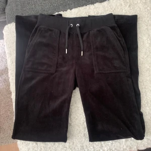 Svarta mjukisbyxor Juicy Couture XS - Svarta mjukisbyxor från Juicy Couture i velour med bred resår och snörning i midjan. Byxorna har stora fickor fram och bak, bakfickorna har lock med silvriga knappar. Perfekta för en chill och trendig look.