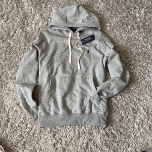Grå hoodie från Polo Ralph Lauren - Snygg grå hoodie från Polo Ralph Lauren med klassisk huva och vit snörning. Tröjan har en broderad blå logga på bröstet, stor ficka framtill och ribbade muddar vid ärmar och nederkant. Perfekt för chill dagar och enkel att matcha med jeans.