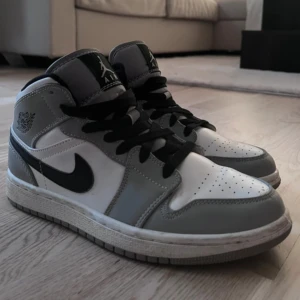 Nike Air Jordan 1 Mid grå/vit - Snygga Nike Air Jordan 1 Mid i grått och vitt med svarta detaljer och klassisk svart swoosh. Skorna har svart snörning, perforerad tå och ikonisk Air Jordan-logga på sidan. Perfekta för dig som gillar streetstyle och vill sticka ut med en tidlös sneaker.