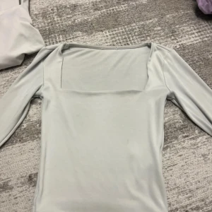 Ljusgrå långärmad topp med fyrkantig ringning - En stilren ljusgrå långärmad topp med snygg fyrkantig ringning. Toppens enkla design gör den lätt att matcha och den har en slim passform som sitter snyggt på kroppen. Perfekt för dig som gillar minimalistisk stil.