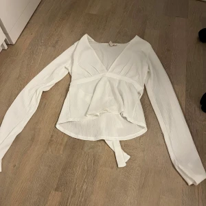 Vit omlottopp från Nelly - Supersnygg vit omlottopp från Nelly med lång ärm och v-ringning. Toppen har knytband i midjan och är tillverkad i ett lätt, ribbat material som ger en skön och trendig look. 