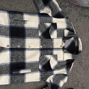 Rutig overshirt i blått och vitt - Snygg overshirt med stora rutor i blått, vitt och grått. Jackan har två bröstfickor med lock och knappar, klassisk krage och knäppning framtill. Perfekt lager-på-lager-plagg för en chill och trendig look.