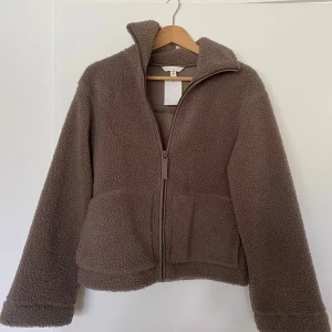 Brun teddyjacka från H&M, stl S - Mysig brun teddyjacka från H&M i storlek S. Jackan har dragkedja framtill, två stora fickor och hög krage. Perfekt för kyliga dagar när du vill vara både varm och snygg. Jackan har en loose passform och är tillverkad i mjuk fleece.