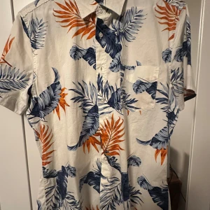 Kortärmad blommig och somrig skjorta i bomull - Snygg kortärmad skjorta med tropiskt blommönster i blått och orange på vit botten. Skjortan har klassisk krage, bröstficka och knäppning framtill. Perfekt för sommarens alla äventyr och chill dagar.