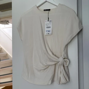 Beige topp med knyt från Zara - Stilren beige topp från Zara med korta ärmar och snygg knytdetalj i sidan. Toppen har rund halsringning och är tillverkad i ett mjukt material som känns skönt mot huden. Perfekt för dig som gillar minimalistisk och trendig stil.