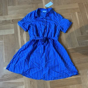 Blå randig kortklänning Holly & Whyte - Snygg blå kortklänning från Holly & Whyte med vita ränder, skjortkrage och knappar framtill. Klänningen har korta ärmar och ett matchande bälte i midjan som knyts till rosett. Tillverkad i mjukt EcoVero-material som känns lätt och fräscht.