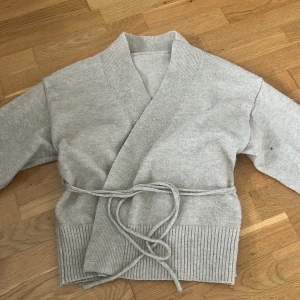 Beige omlottkofta med knytband - Mysig grå kofta i omlottmodell med bred ribbad kant och knytband i midjan. Perfekt för lager-på-lager och chill dagar. Tröjan har lång ärm och är stickad i mjukt material som känns skönt mot huden. Använd fåtal gånger 