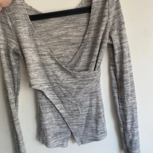 Grå omlottopp med v-ringning - Snygg grå omlottopp med v-ringning och lång ärm. Toppen har ett ribbat mönster och en cool omlottdetalj framtill som ger en trendig look. Perfekt att styla med jeans eller kjol för en chill vibe.