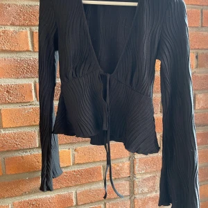 Svart ribbad omlottkofta Gina Tricot - Svart omlottkofta från Gina Tricot i storlek S. Tröjan har ribbad struktur, långa ärmar och knytband framtill. Den har en lätt croppad passform och är tillverkad i mjukt syntetmaterial. Perfekt för dig som gillar stilrena och trendiga plagg.