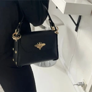 Svart handväska med gulddetaljer - Snygg svart handväska i skinn med guldfärgad kedja och coolt bi i metall framtill. Väskan har både handtag och axelrem i kedja, samt dekorativa nitar på sidorna. Perfekt storlek för det viktigaste och ger en lyxig vibe till din outfit.En snygg liten väska. Storlek 20x13. Aldrig använt så super fint skick! Köptes för 700kr! 