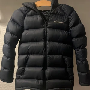 Mörk blå dunjacka Peak Performance - Snygg svart dunjacka från Peak Performance med huva och diskret logga på bröstet. Jackan är fluffig och har dragkedja framtill, perfekt för kalla dagar. Klassisk pufferstil med lång ärm och enkel design som funkar till allt.