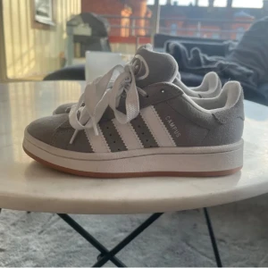 Grå Adidas Campus sneakers  - Snygga Adidas Campus sneakers i grå mocka med klassiska vita ränder och vit sula. Skorna har rund tå, platt sula och snörning. Perfekta för dig som gillar en clean och stil. Skriv för fler bilder