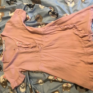 Ljusrosa kort klänning med volang (barnkläder) - Supersöt ljusrosa klänning från 157 med korta puffärmar, knappar framtill och volangdetaljer längs kjolkanten. Mjuk och lätt i materialet, perfekt för dig som gillar en feminin och chill stil. Passar dig som vill ha något enkelt men ändå gulligt.