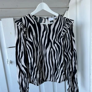 Zebramönstrad blus från VILA - Säljer en svartvit blus från VILA med zebramönster. Blusen har långa ärmar, volangdetaljer vid axlarna och rund hals. Materialet är mjukt och lätt, perfekt för dig som gillar att sticka ut med coola mönster. Passar dig som vill ha en trendig look.