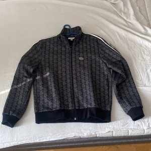 Svart mönstrad track jacket från Lacoste  - Snygg svart track jacket från Lacoste med geometriskt mönster över hela jackan och klassiska vita ränder längs ärmarna. Dragkedja framtill, ribbade muddar och ståkrage. Ikoniska gröna krokodilloggan på bröstet. Perfekt för en sportig och stilren look. Liten i storleken passar dig som har medium i vanliga fall 