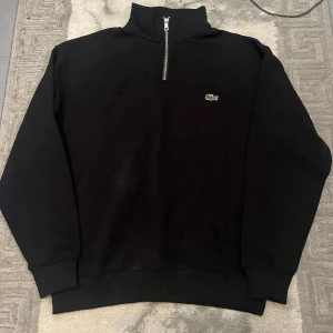 Svart half zip tröja från Lacoste - Svart långärmad tröja från Lacoste med half zip och klassisk krokodillogga på bröstet. Hög krage och ribbade muddar ger en clean och sportig look. Perfekt för dig som gillar stilrena och enkla plagg med premiumkänsla.